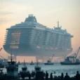 Editorial photograph of a colossal conceptual floating city vessel at dawn, viewed from a working harbor.
