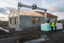 Editorial photograph of a low-rise housing construction site in Denmark, a gantry-mounted concrete printer spanning.