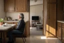 Editorial photograph inside a modest American manufactured home, a resident seated at a kitchen table beside.