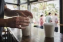 Editorial photograph inside a small Florida coffee shop at morning rush, a barista’s hands folding.