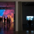 Editorial photograph inside a contemporary art museum, looking across a deep doorway between an immersive projection.