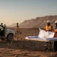 An illustrative editorial photograph in Saudi Arabia’s northwestern desert: two infrastructure surveyors beside a folding table.