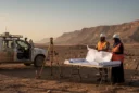 An illustrative editorial photograph in Saudi Arabia’s northwestern desert: two infrastructure surveyors beside a folding table.