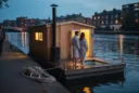 Editorial photograph of a modest floating, wood-fired sauna on a London waterway at blue hour, viewed.