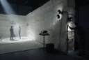 Editorial photograph of an experimental mycelium-module pavilion in a dark exhibition hall, two visitors crossing faint.