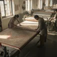 Editorial photograph inside a rug-making workshop in Rangpur, Bangladesh, showing workers examining a large woven rug.