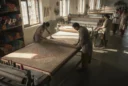 Editorial photograph inside a rug-making workshop in Rangpur, Bangladesh, showing workers examining a large woven rug.