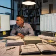 Editorial photograph inside a working architecture studio at dusk: an architect studies a BIM wall assembly.