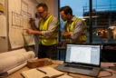 Editorial photograph inside a working British architecture studio at dusk: an architect and contractor debate.