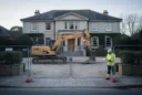 Editorial photograph of a large, apparently well-maintained mansion behind temporary construction fencing, a tracked excavator parked.